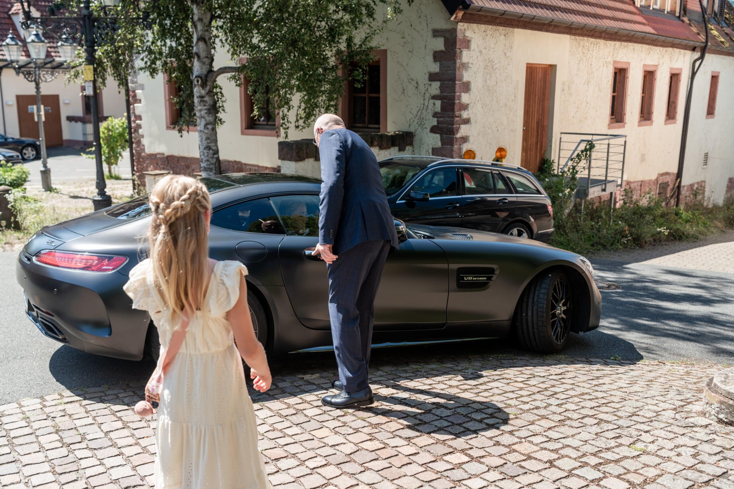 Brautmädchen und Mann am Auto auf Kopfsteinpflaster.