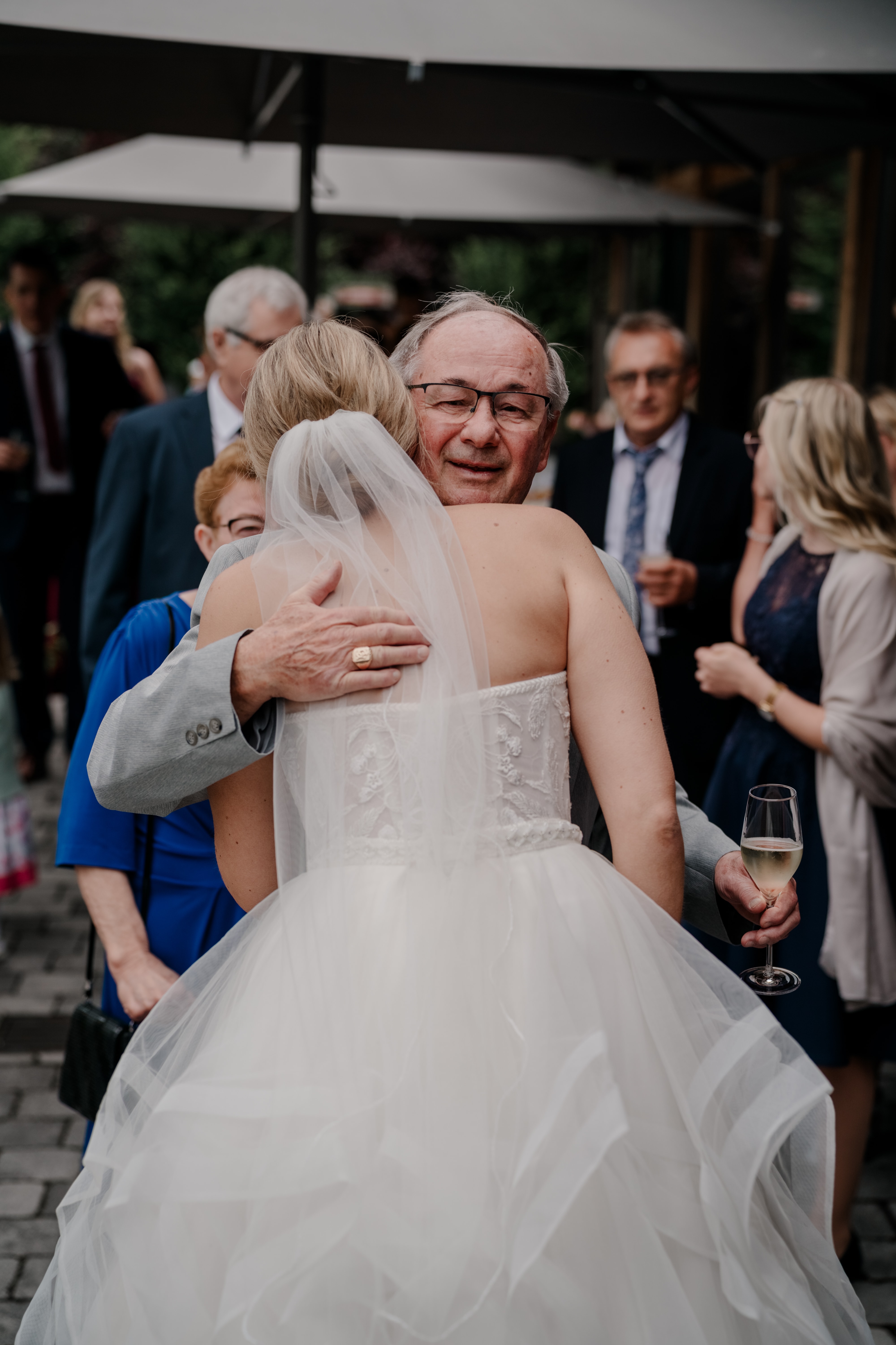 Braut umarmt älteren Gast, Sektglas in der Hand.