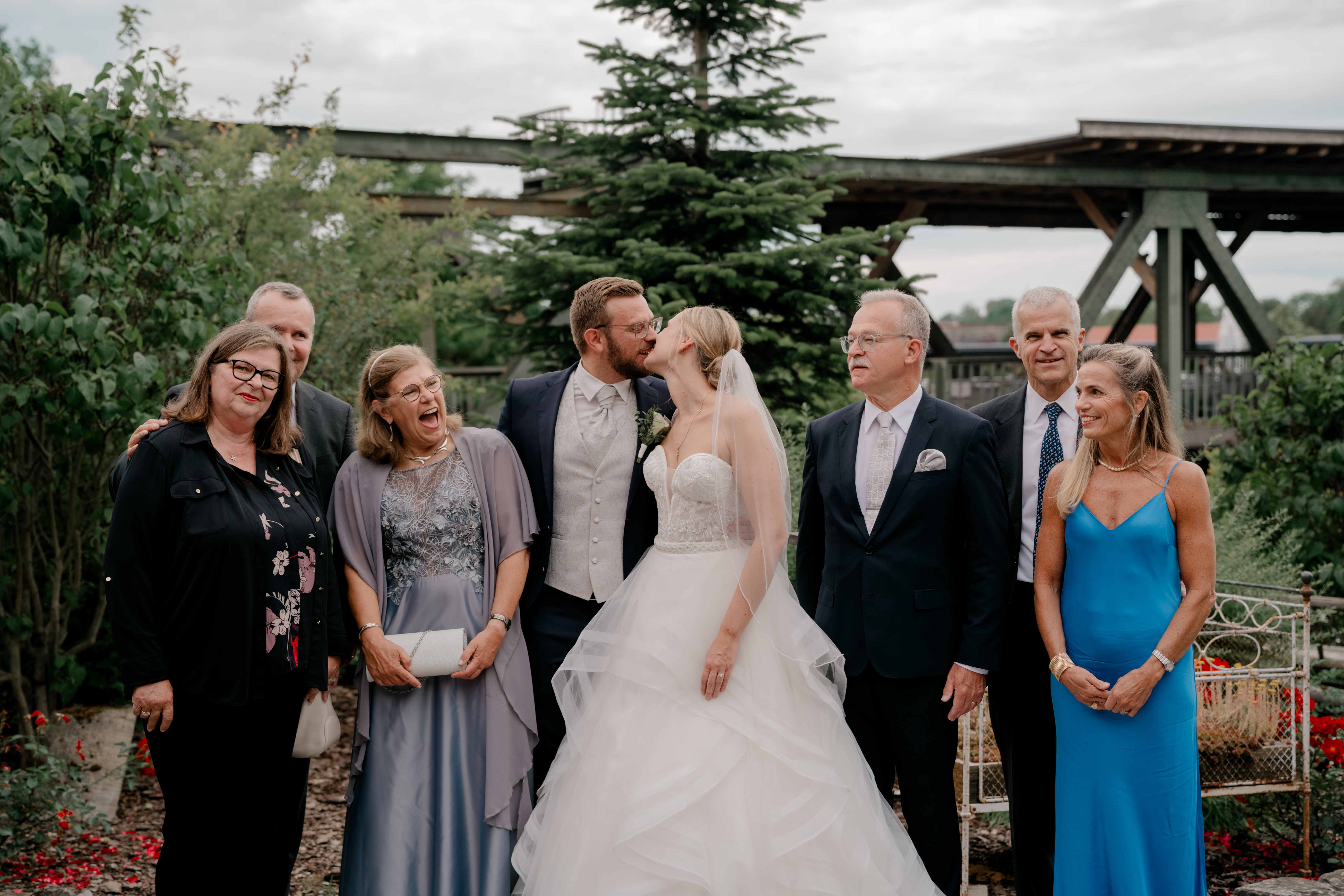 Brautpaar küsst sich mit Gästen im Garten.