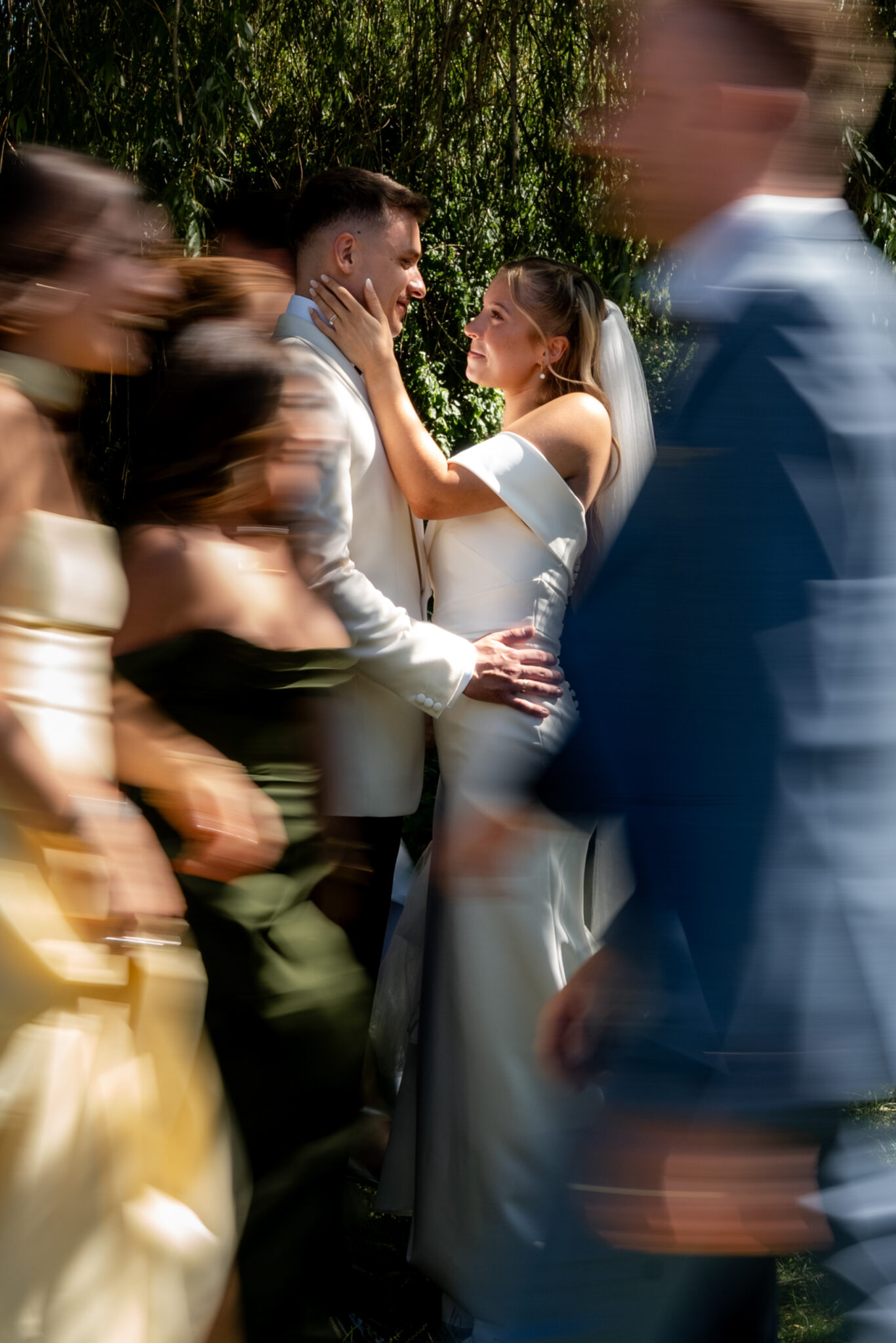 Brautpaar umarmt sich im Garten, Gäste ziehen vorbei