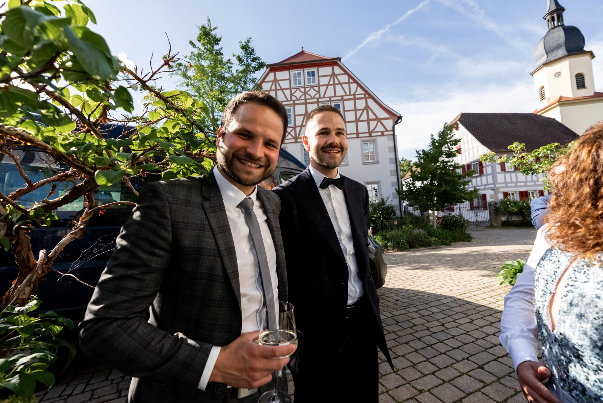 Hochzeitsgäste im Anzug mit Sekt vor Fachwerk und Kirche