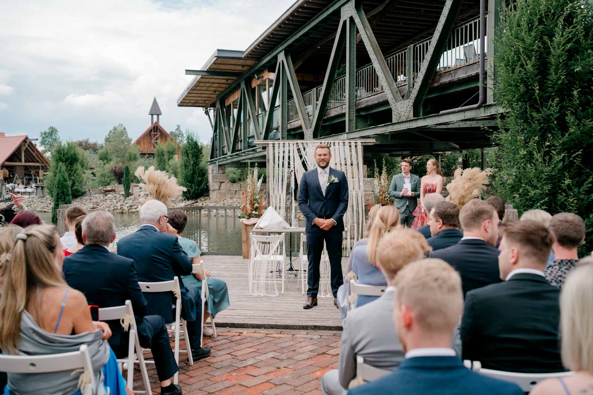 Eine Hochzeitszeremonie im Freien, Gäste sitzen auf Stühlen vor einem Bräutigam, der vor einem dekorativen Hintergrund steht. Hochzeit Brückenbaron Freie Trauung