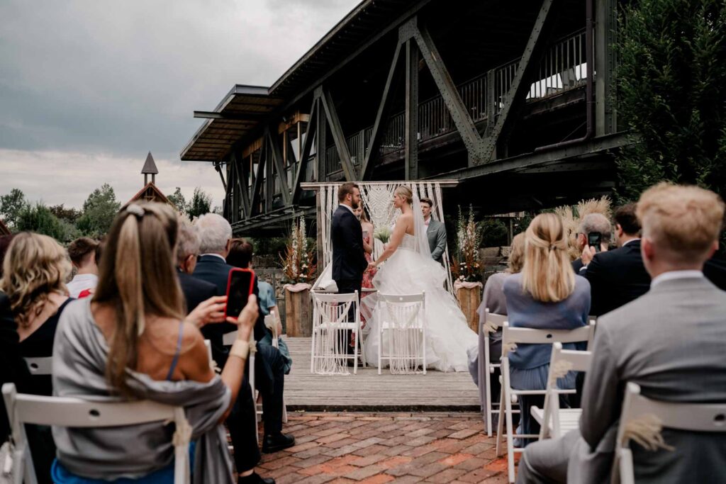 Hochzeitszeremonie im Freien, bei der ein Brautpaar vor einem dekorierten Altar steht, umgeben von sitzenden Gästen. Hochzeit Brückenbaron Freie Trauung