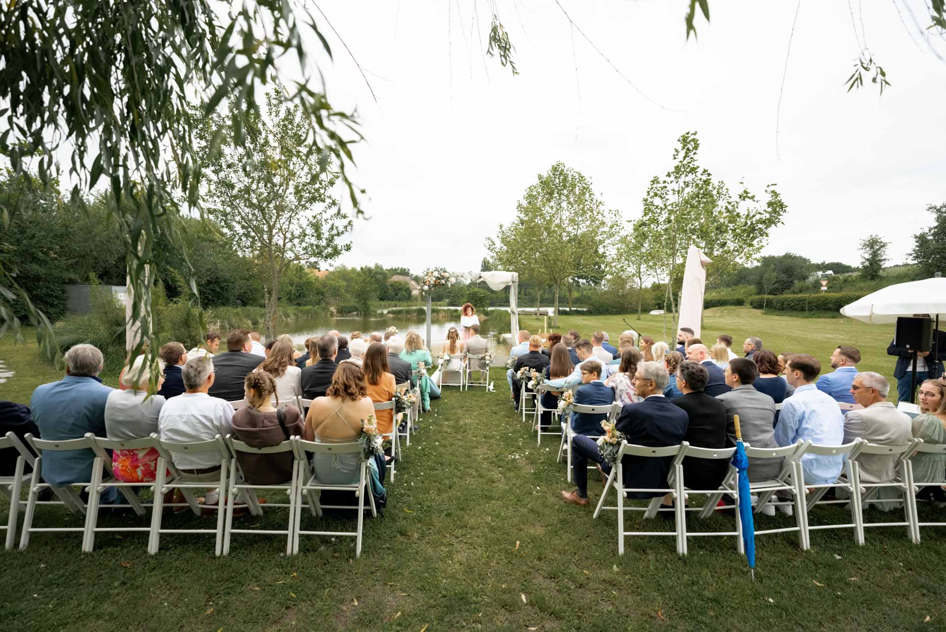 Hochzeitszeremonie im Freien mit vielen Gästen, die auf weißen Klappstühlen vor einem Teich sitzen. Im Hintergrund stehen Bäume und eine Person vorne liest aus einem Buch unter einem Blumenbogen.