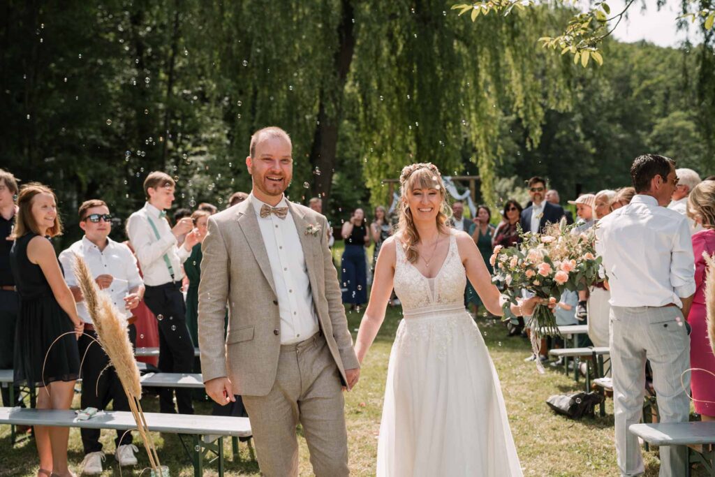 Braut und Bräutigam lächeln, während sie bei einer Hochzeit draußen zwischen den Gästen hindurchgehen.