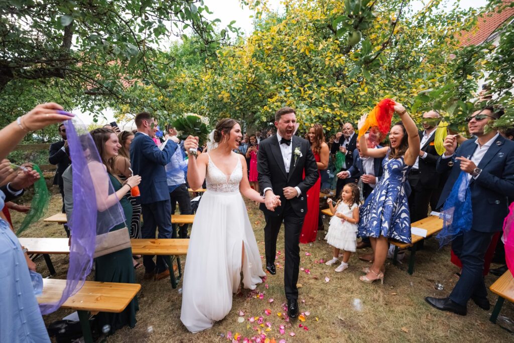 Ein Hochzeitspaar geht durch eine fröhliche Menschenmenge im Freien, umgeben von lachenden Gästen, die bunte Tücher schwenken.