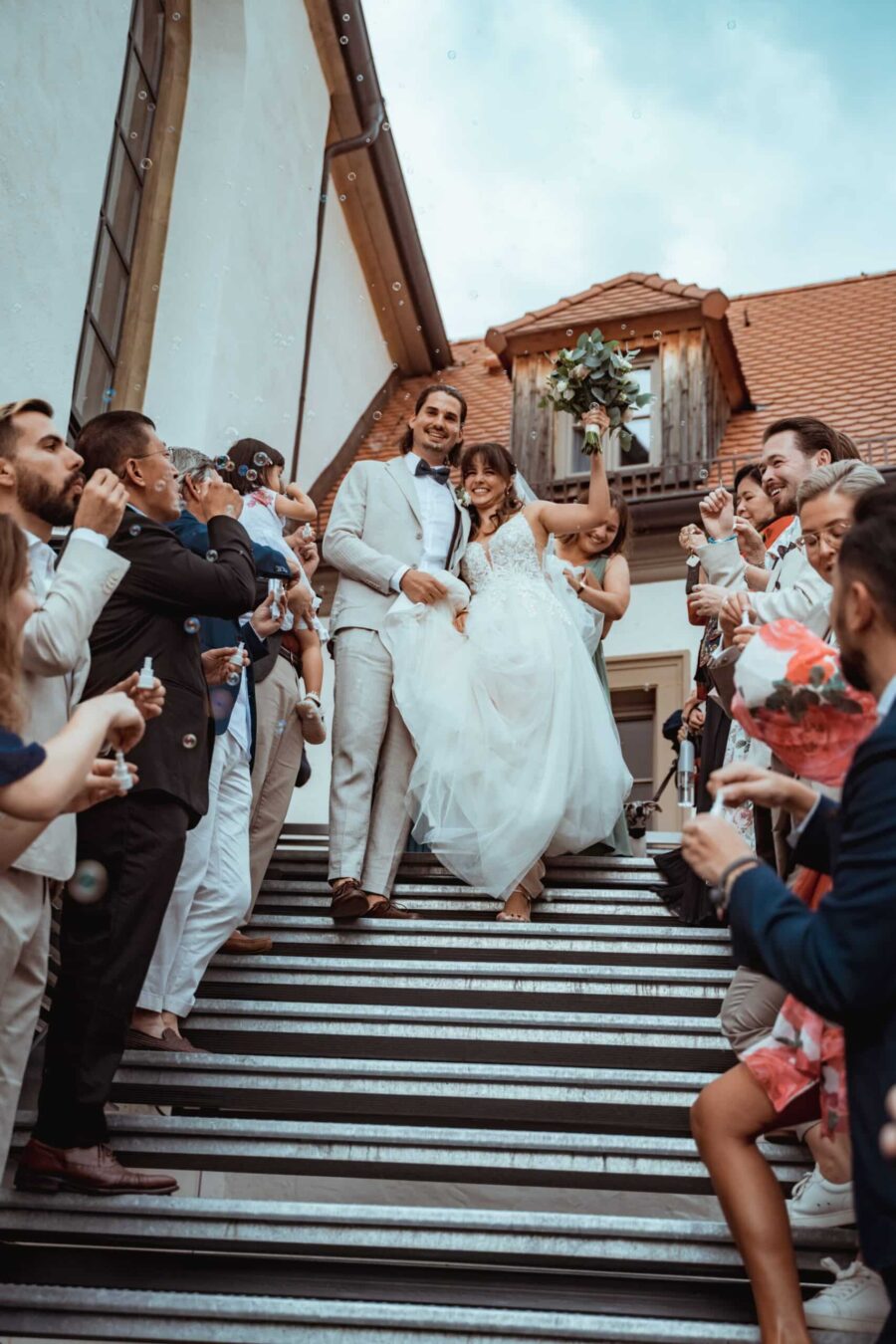 Brautpaar auf Treppe, umgeben von feiernden Gästen.