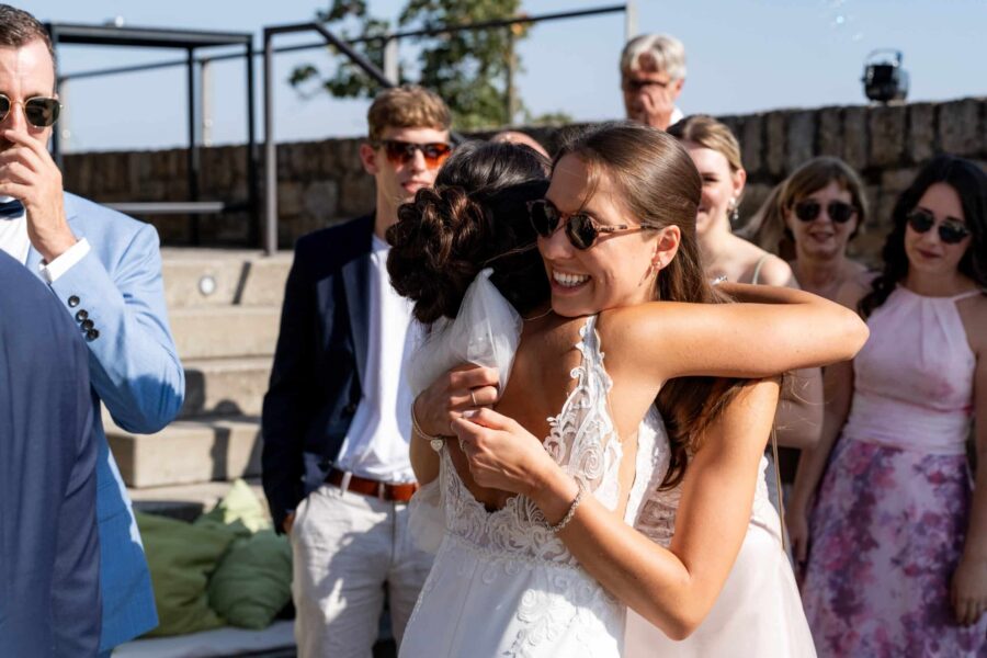 Braut umarmt Freundin bei sonniger Hochzeitsfeier draußen.