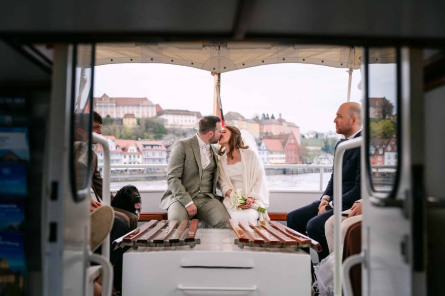 Hochzeit Insel Mainau Bodensee – Heiraten am Bodensee 128 Brautpaarkuss auf dem Boot mit Gästen und Stadtblick