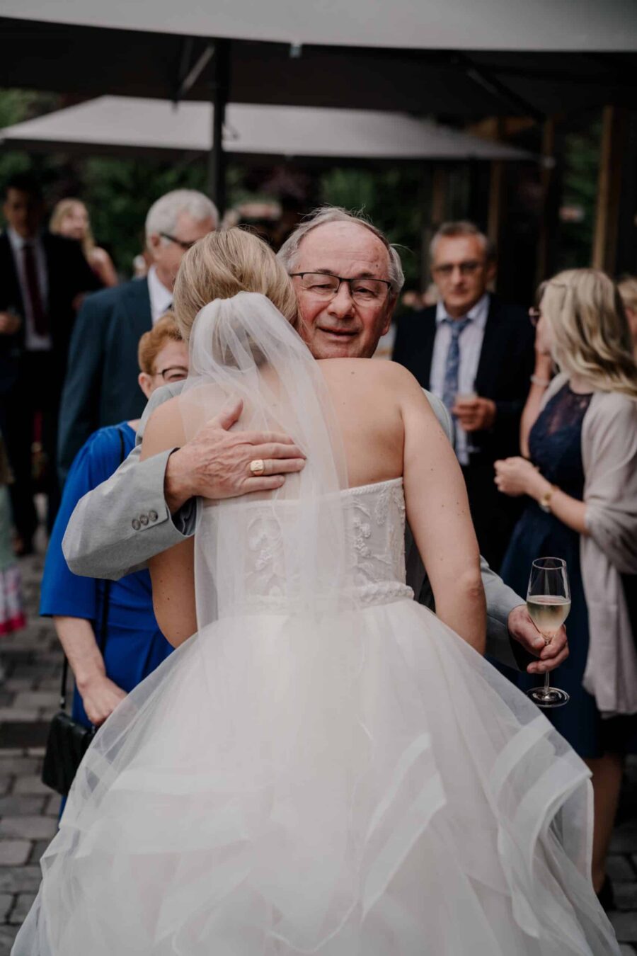 Braut umarmt älteren Gast, Sektglas in der Hand.