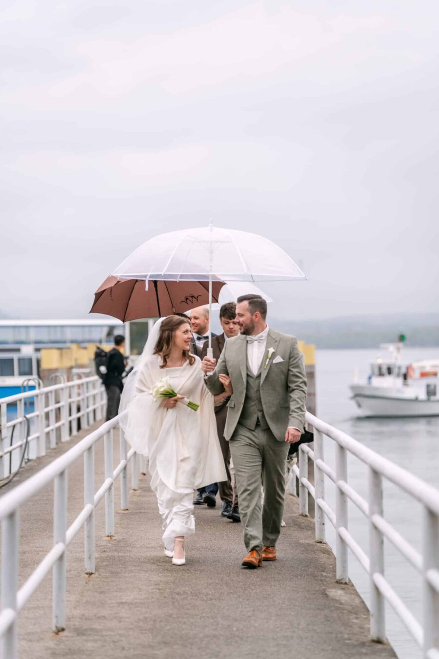 Brautpaar und Gäste mit Schirmen im Regen am See-Steg
