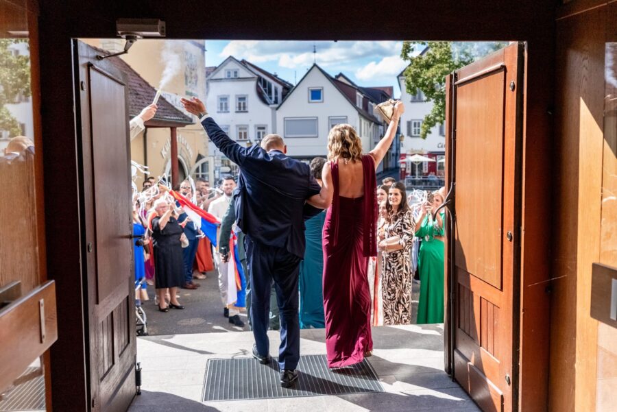 Hochzeitsgäste begrüßen Brautpaar vor Kirche.