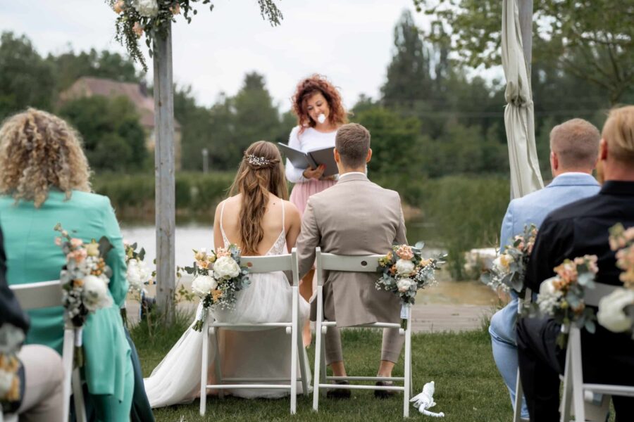 Hochzeitszeremonie am See mit Brautpaar und Gästen.
