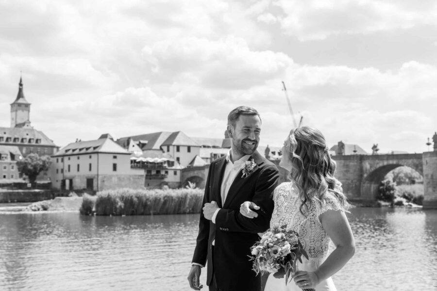 Brautpaar spaziert lachend am Flussufer im Freien.