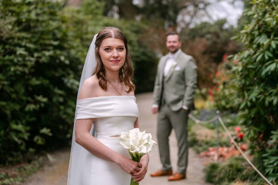 Hochzeit Insel Mainau Bodensee – Heiraten am Bodensee 151 Braut mit Calla-Strauß, Bräutigam unscharf im Garten