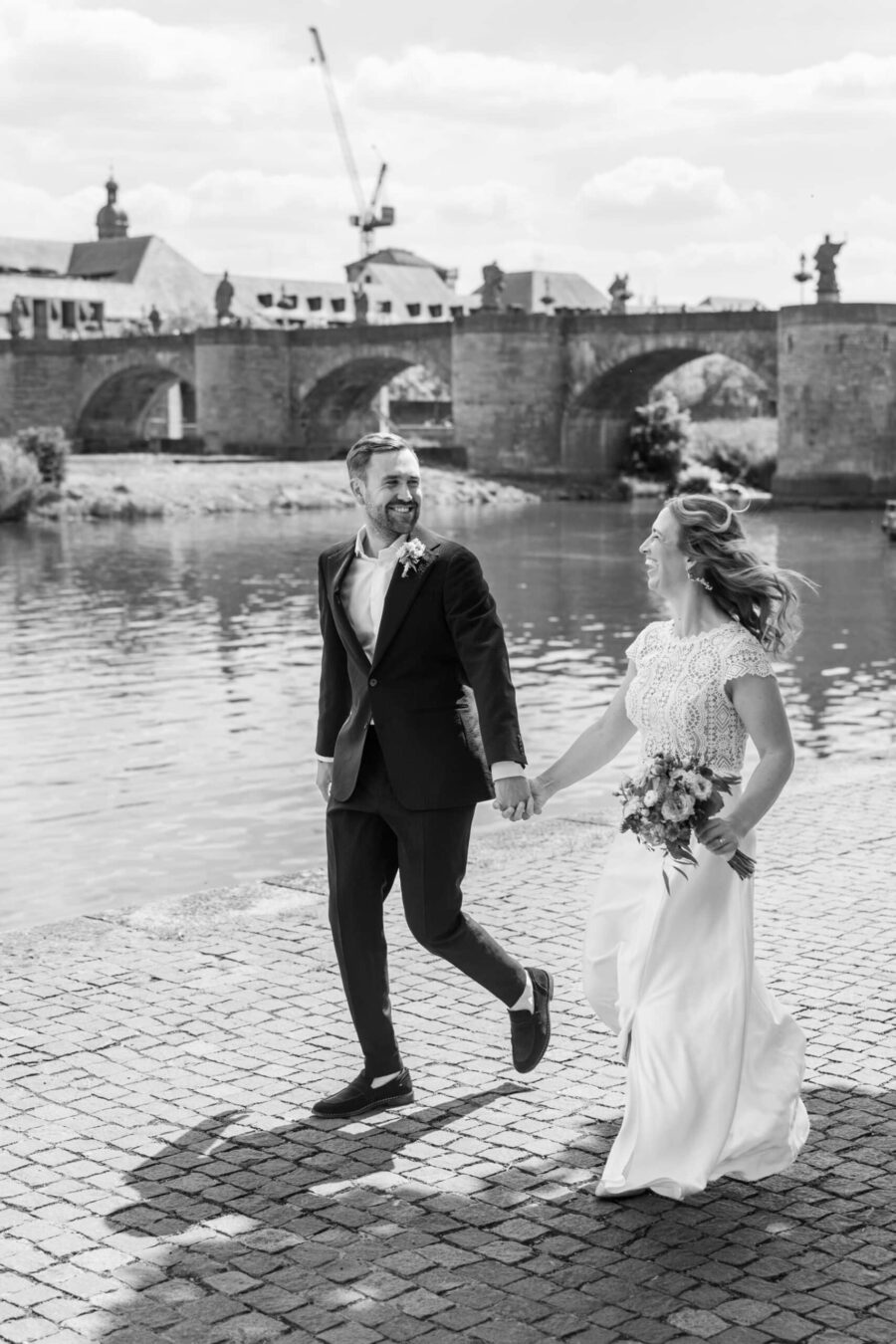 Hochzeit Steinburg Würzburg | Editorial Style & Party | Florian Knusper 19 Brautpaar spaziert am Flussufer vor historischer Brücke