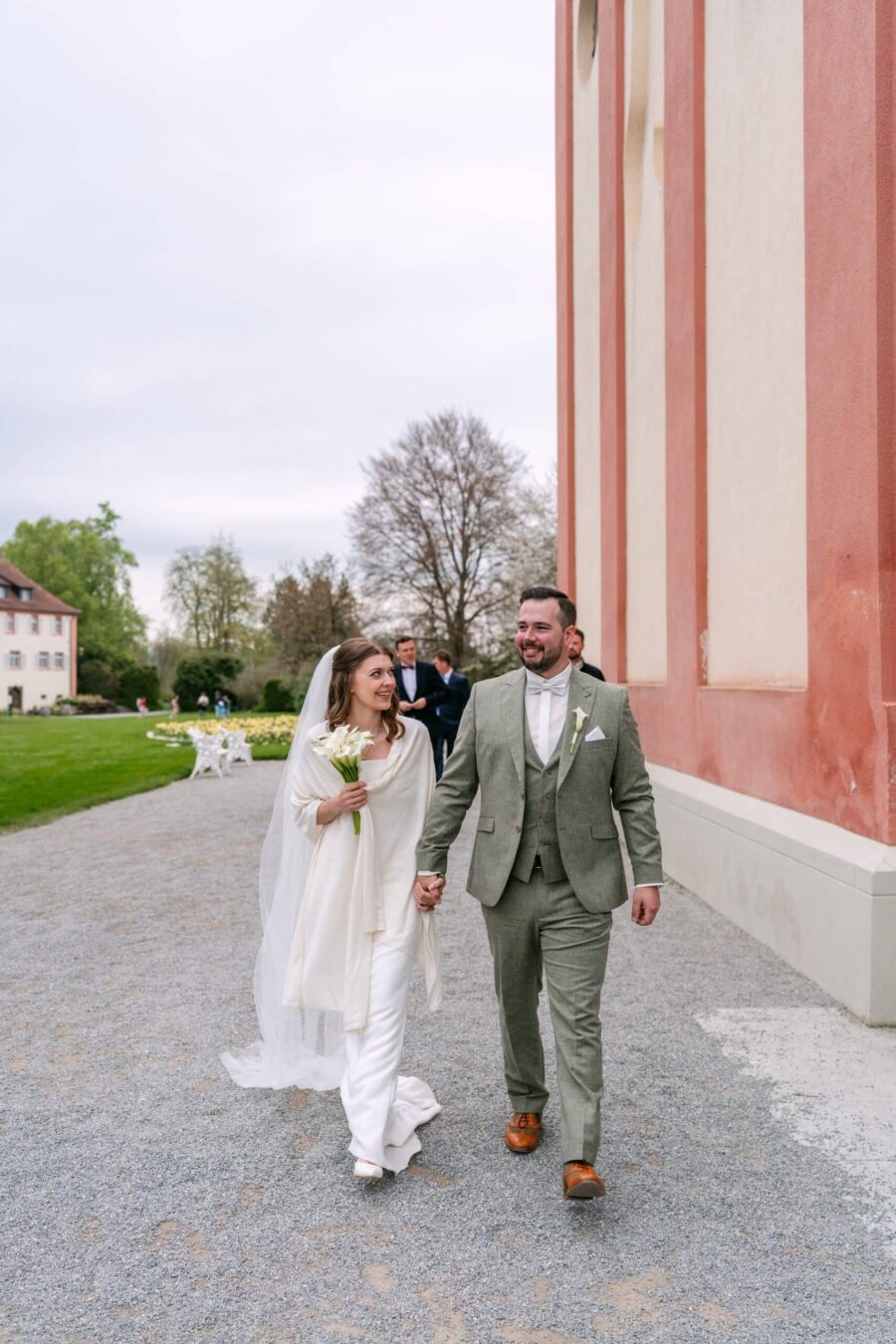 Hochzeit Insel Mainau Bodensee – Heiraten am Bodensee 209 Brautpaar spaziert Hand in Hand vor historischer Fassade