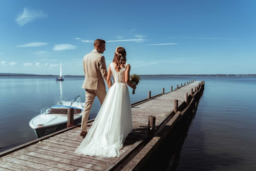 Brautpaar auf Steg am See mit Segelboot im Hintergrund
