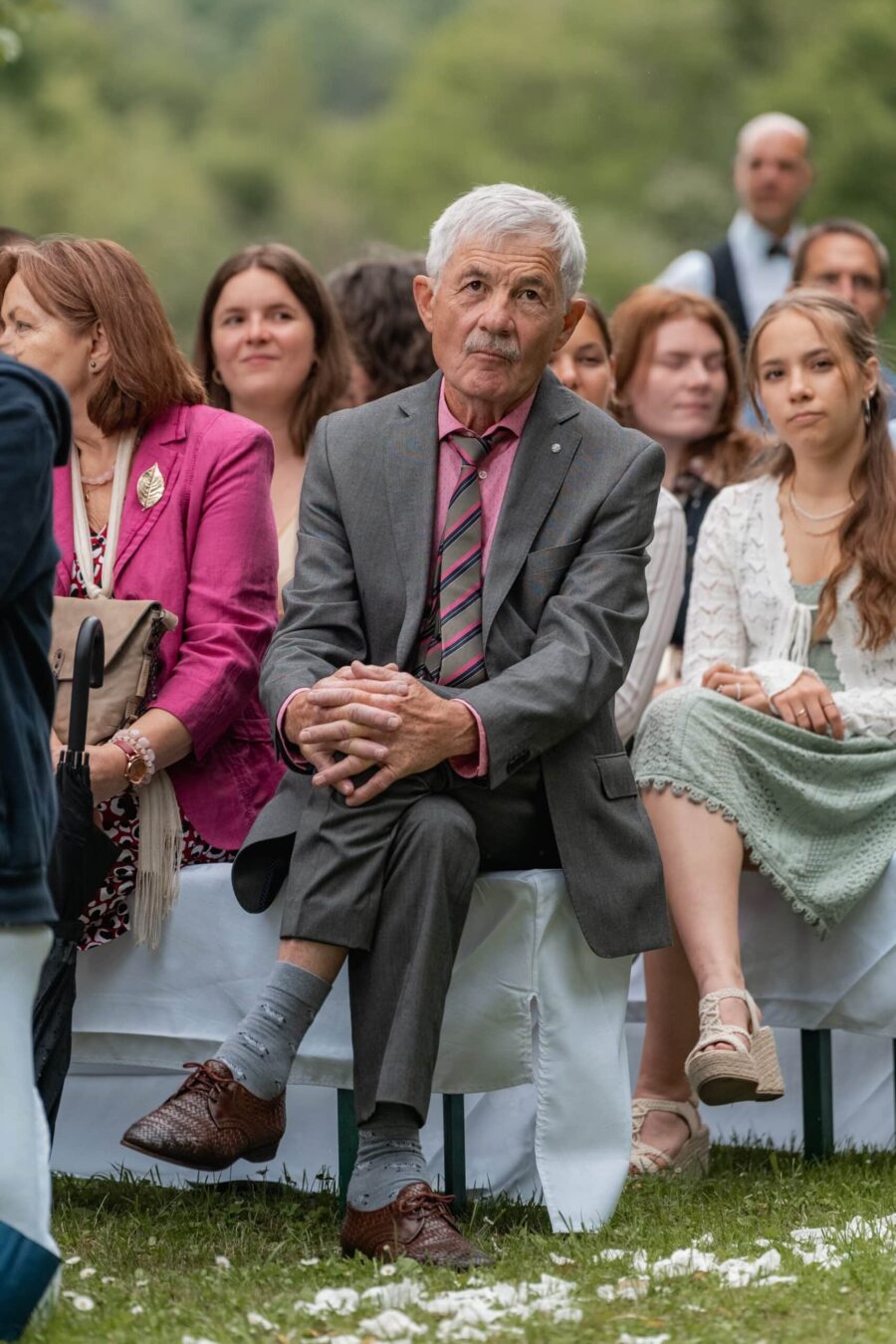 Hochzeitsgäste sitzen im Freien auf weißen Bänken.