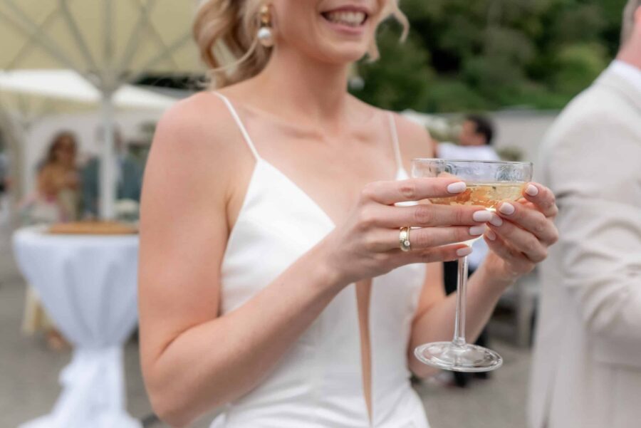 Hochzeit Villa Oriental | Editorial Summer Vibes & Live Cooking 408 Braut mit Sektglas im Freien bei Hochzeitsempfang
