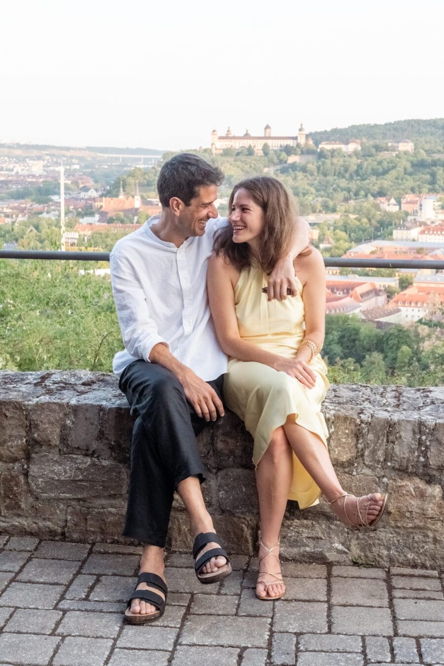 Hochzeit Steinburg Würzburg | Editorial Style & Party | Florian Knusper 68 Paar sitzt lächelnd auf Steinmauer mit Stadtpanorama.