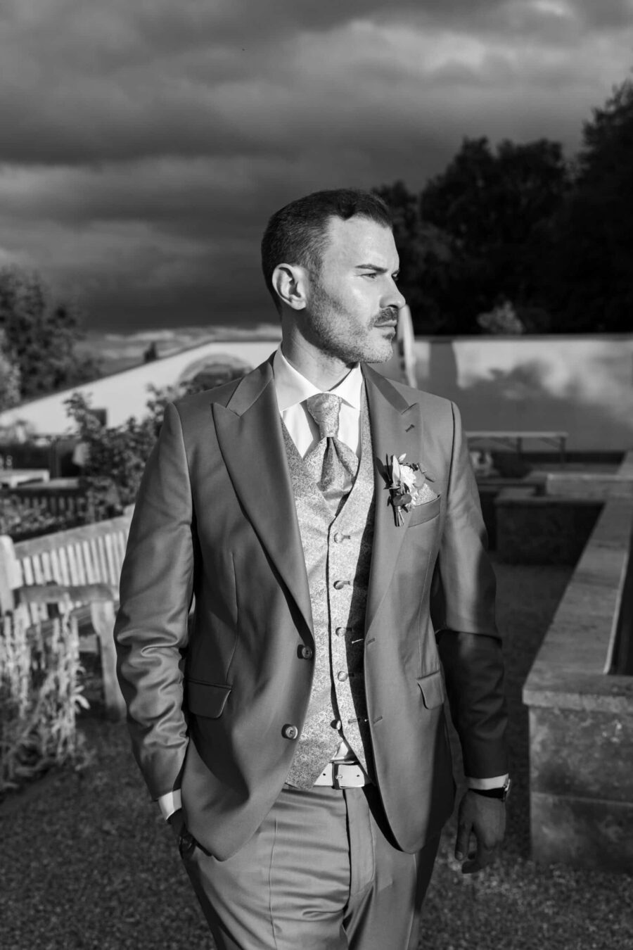 Mann im Anzug in einem Garten bei bewölktem Himmel.